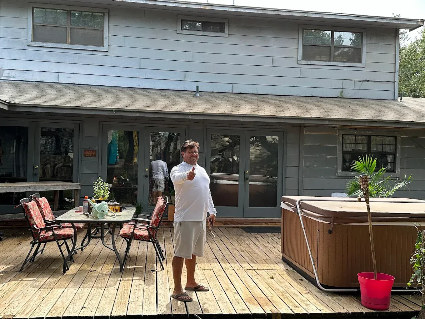 Person standing on a wooden deck outside a house, with patio furniture and a hot tub nearby.