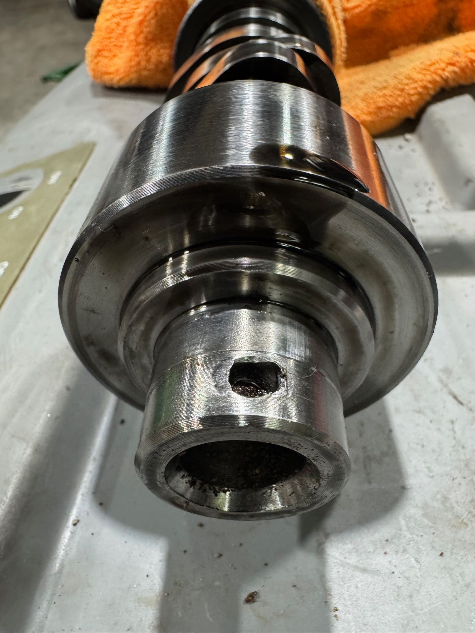 Close-up of a metal camshaft with various lobes and a hole, resting on a gray surface with an orange cloth in the background. | Ace-High Diesel