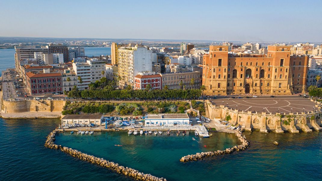 Porto costiero con acque cristalline, frangiflutti in pietra, imbarcazioni da diporto ed edifici storici lungo il lungomare.