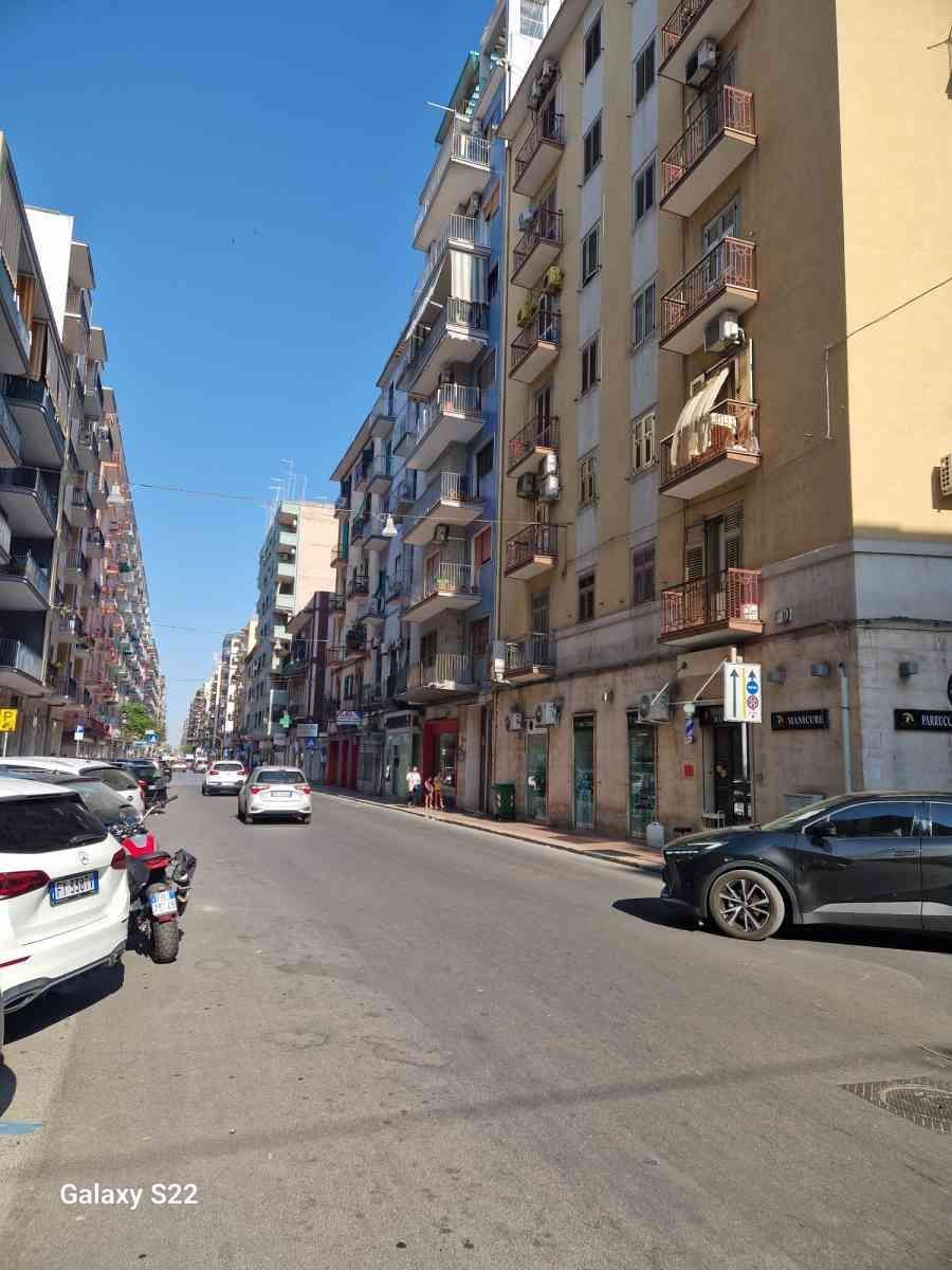 Una soleggiata strada cittadina con palazzi, auto parcheggiate e alcuni veicoli in transito.