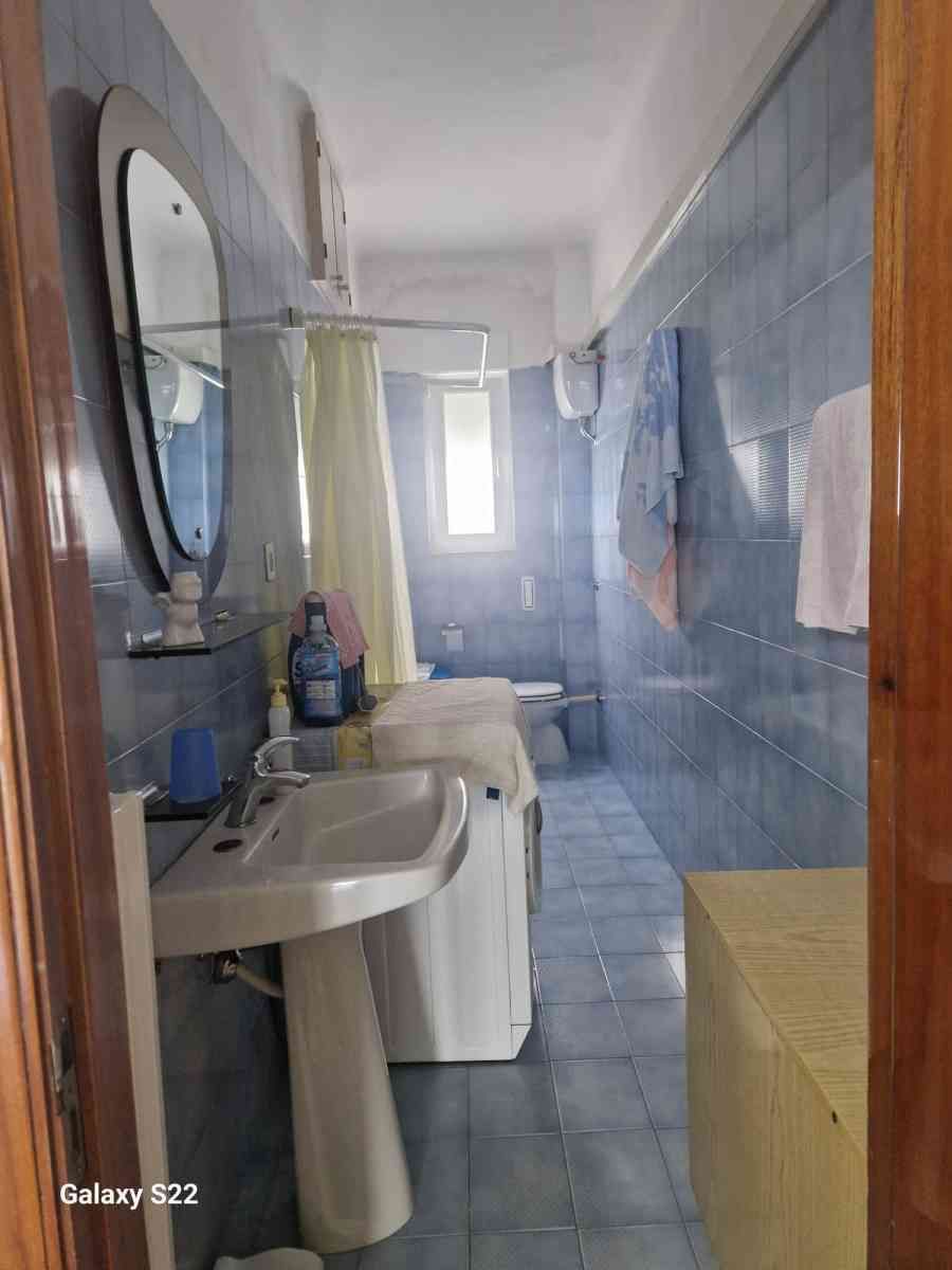 Bagno stretto con piastrelle blu, lavabo, specchio, lavatrice, tenda da doccia e finestra.