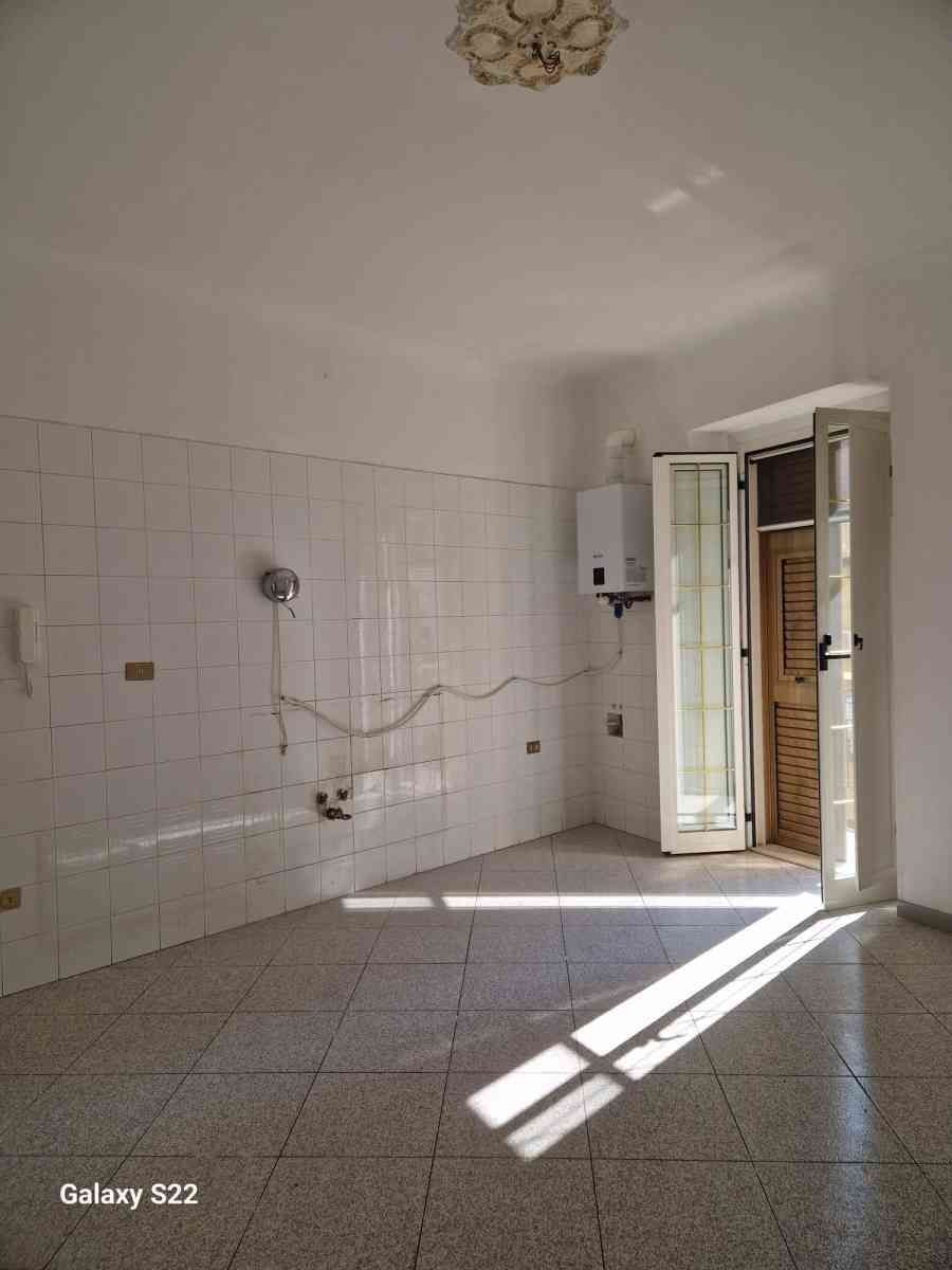 Stanza vuota con pavimento piastrellato di bianco, illuminata dalla luce del sole e con una porta aperta che dà su un balcone.