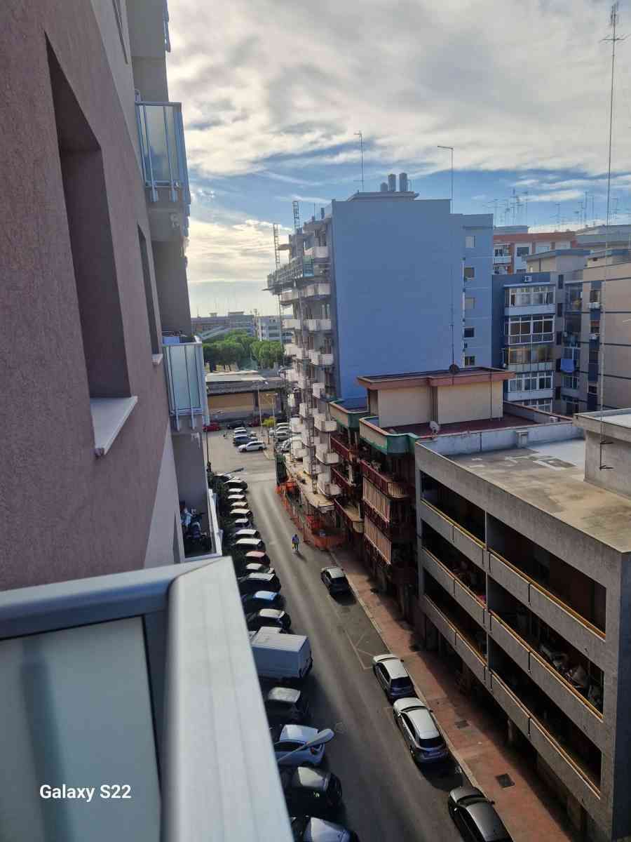 Stretta via cittadina tra palazzi residenziali, con auto parcheggiate e un cielo nuvoloso sullo sfondo.