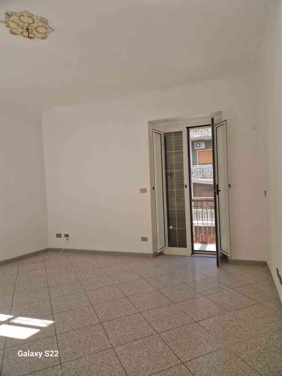 Stanza bianca vuota con pavimento piastrellato e porta-finestra del balcone aperta che lascia entrare la luce naturale.