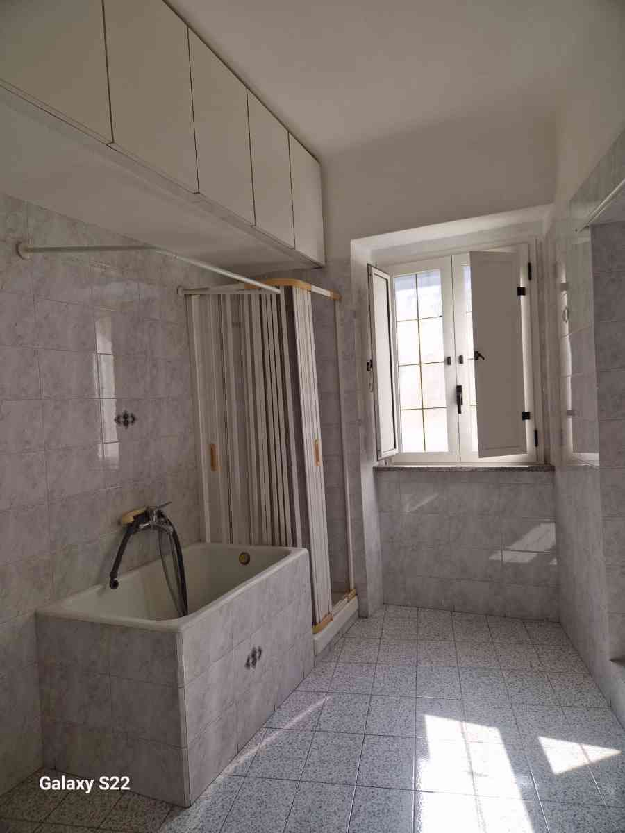 Bagno luminoso e bianco con vasca, tenda da doccia, armadietti e finestra che lascia entrare la luce del sole.