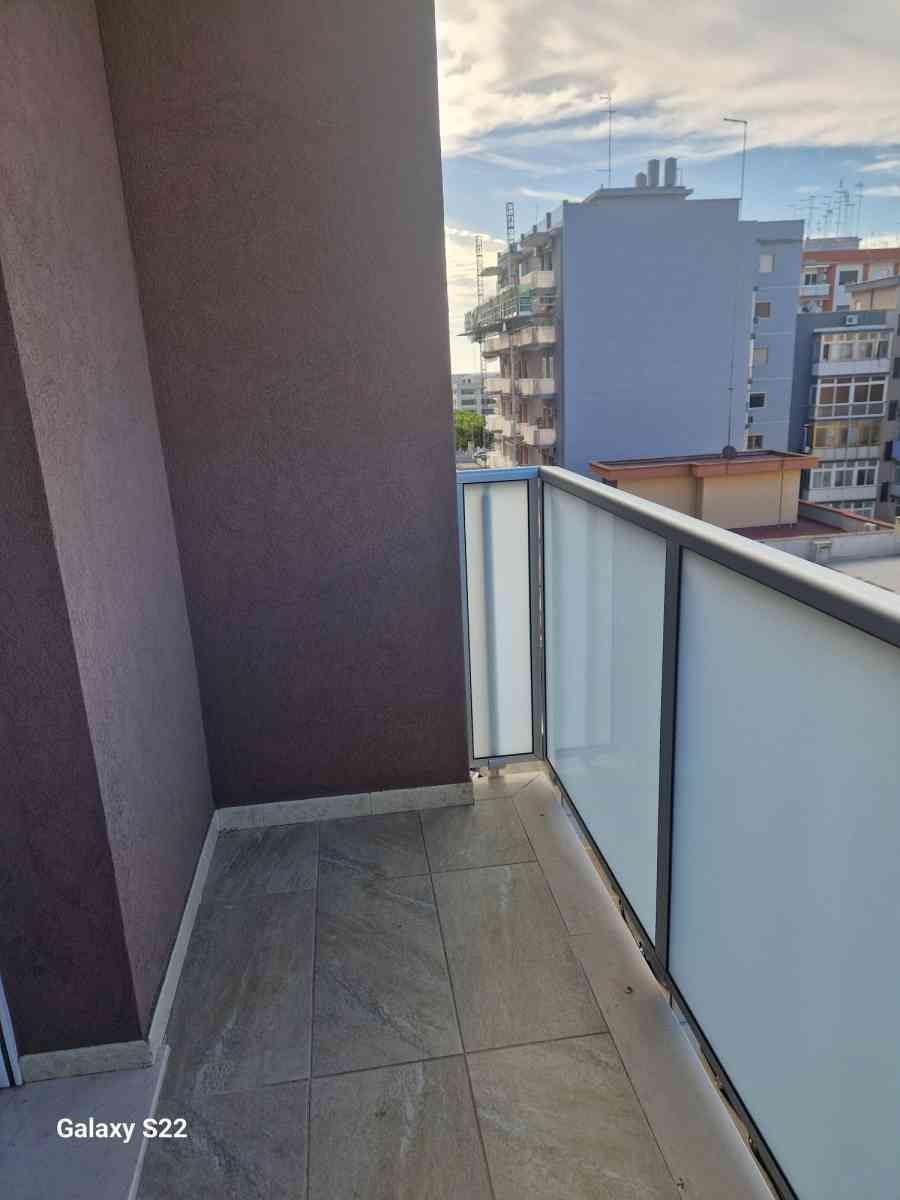 Stretto balcone di un appartamento con piastrelle grigie, ringhiera in vetro satinato e vista sulla città in lontananza.