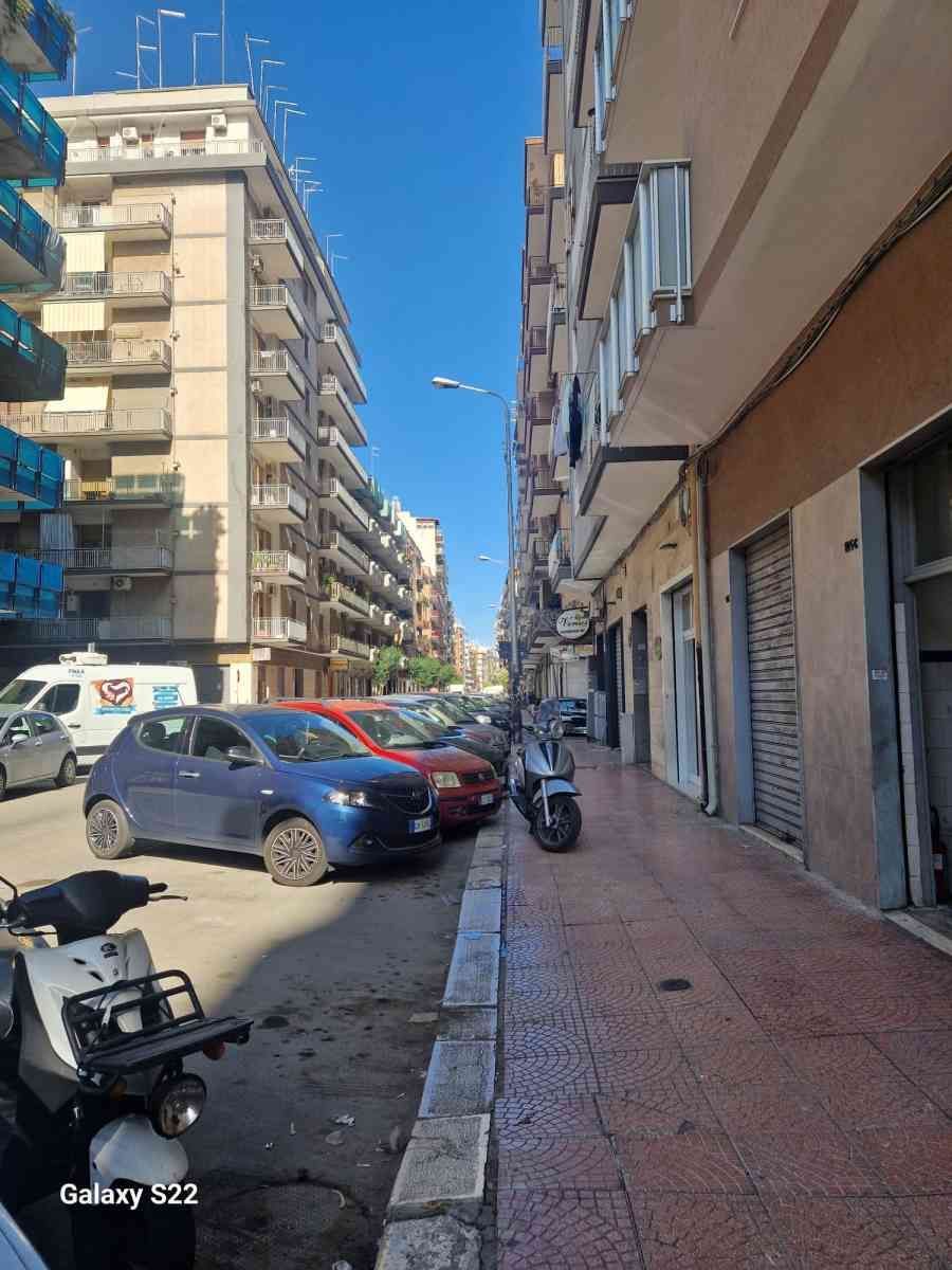 Stretta via cittadina con auto parcheggiate, palazzi e un cielo azzurro limpido.