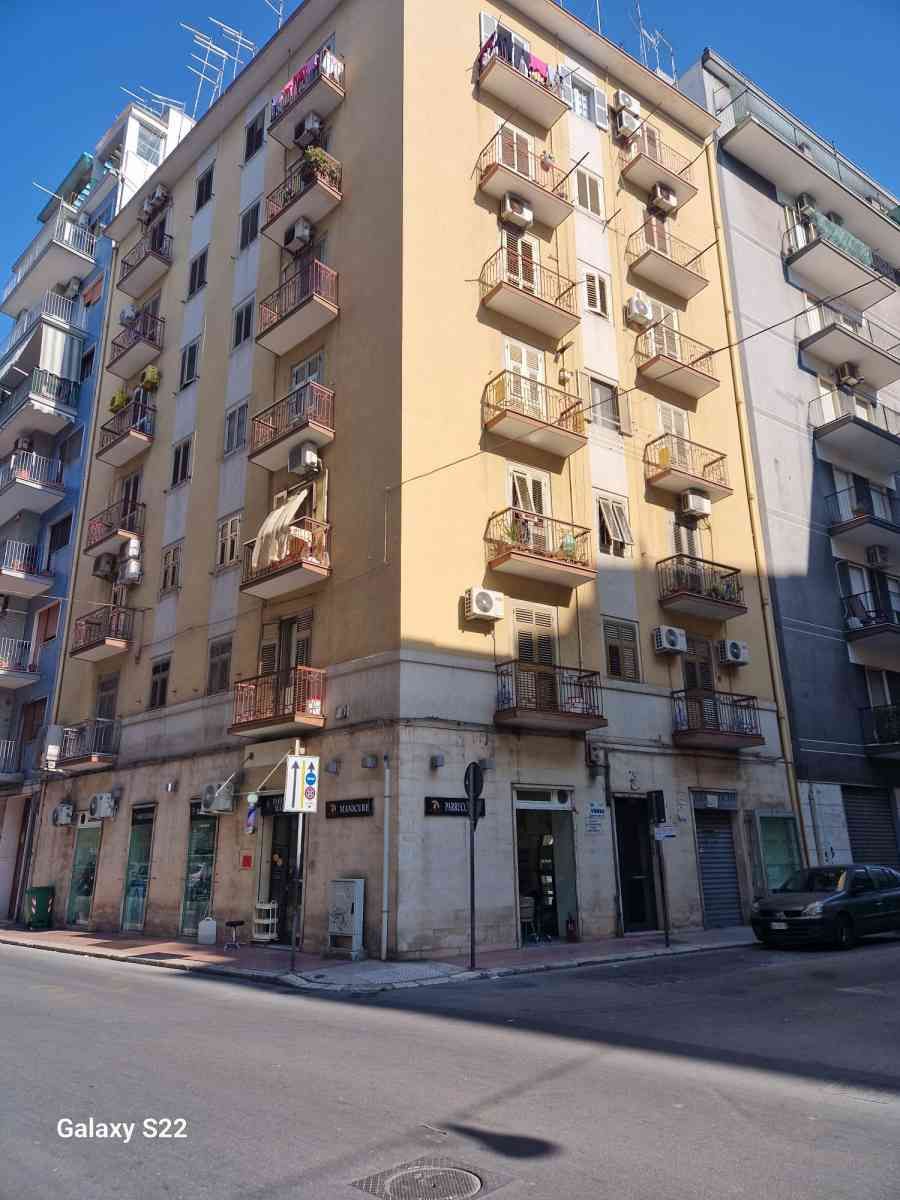 Angolo di strada soleggiato con un palazzo beige, balconi, vetrine e auto parcheggiate.
