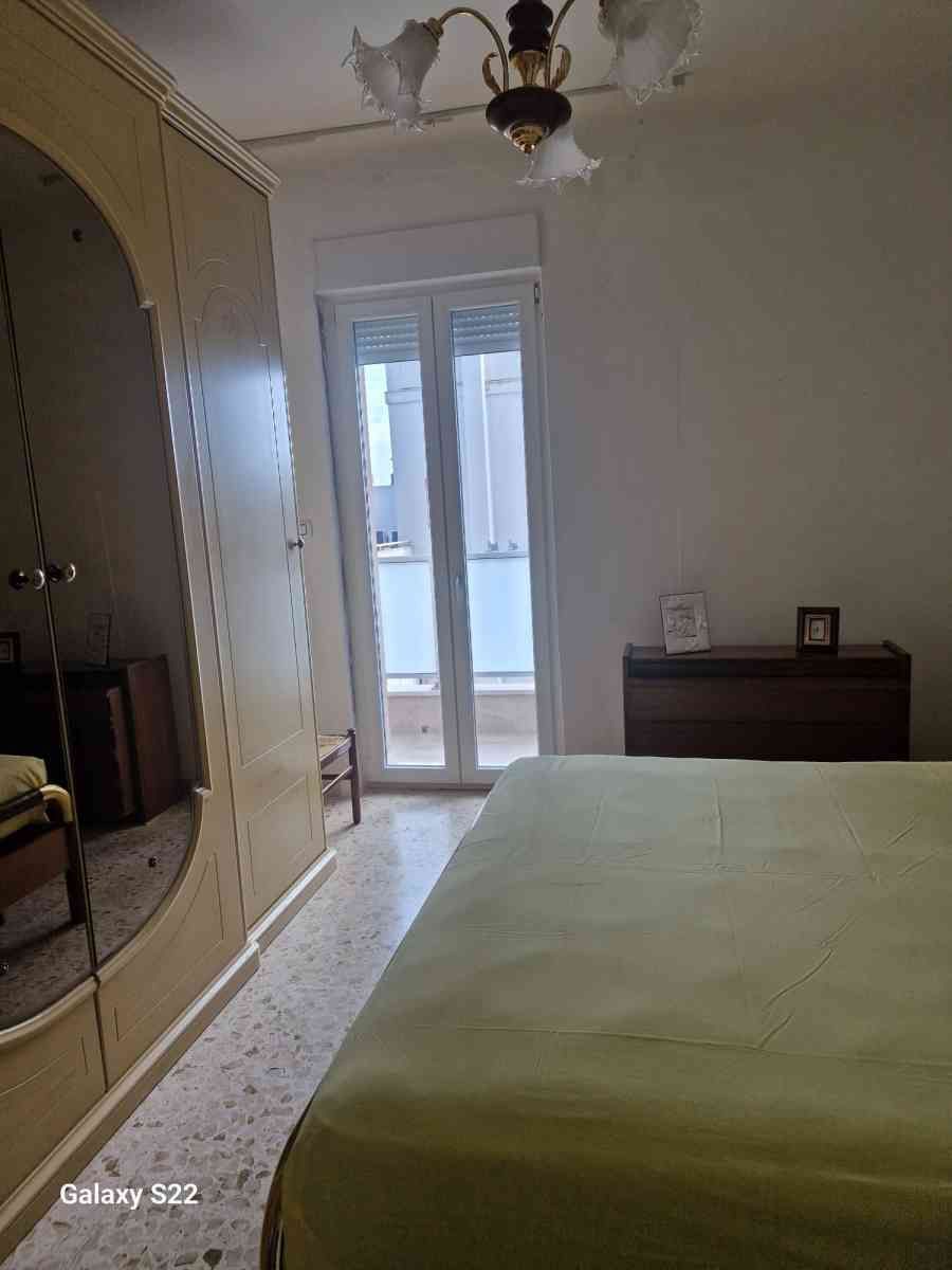 Piccola camera da letto con letto, armadio con specchio, cassettiera e porte finestre che danno su un balcone.