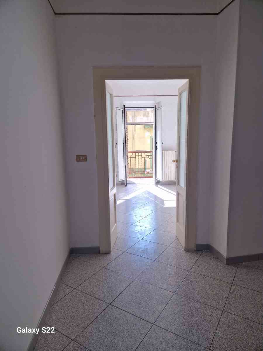 Corridoio vuoto con pavimento piastrellato che conduce a una porta luminosa e a un balcone.