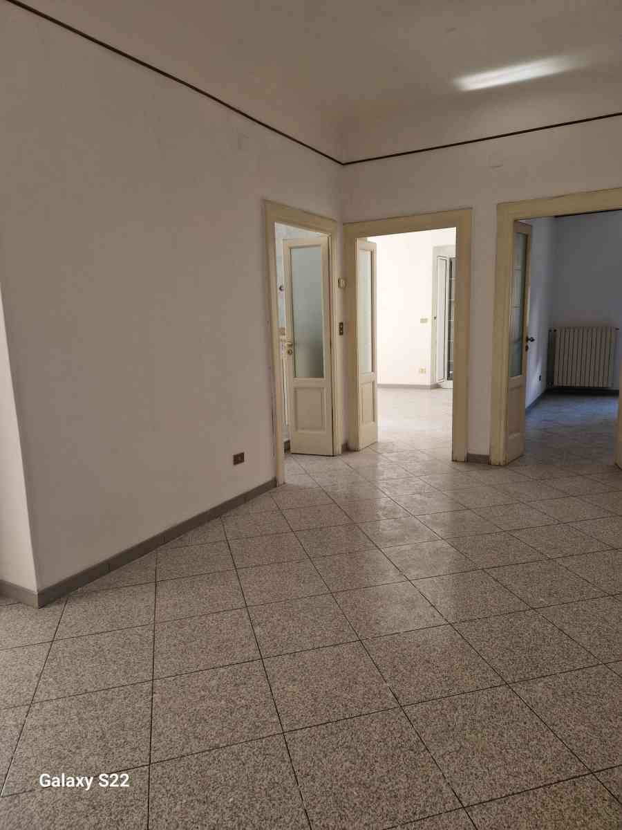 Stanza vuota piastrellata con pareti bianche e porte aperte che conducono a stanze adiacenti più luminose.