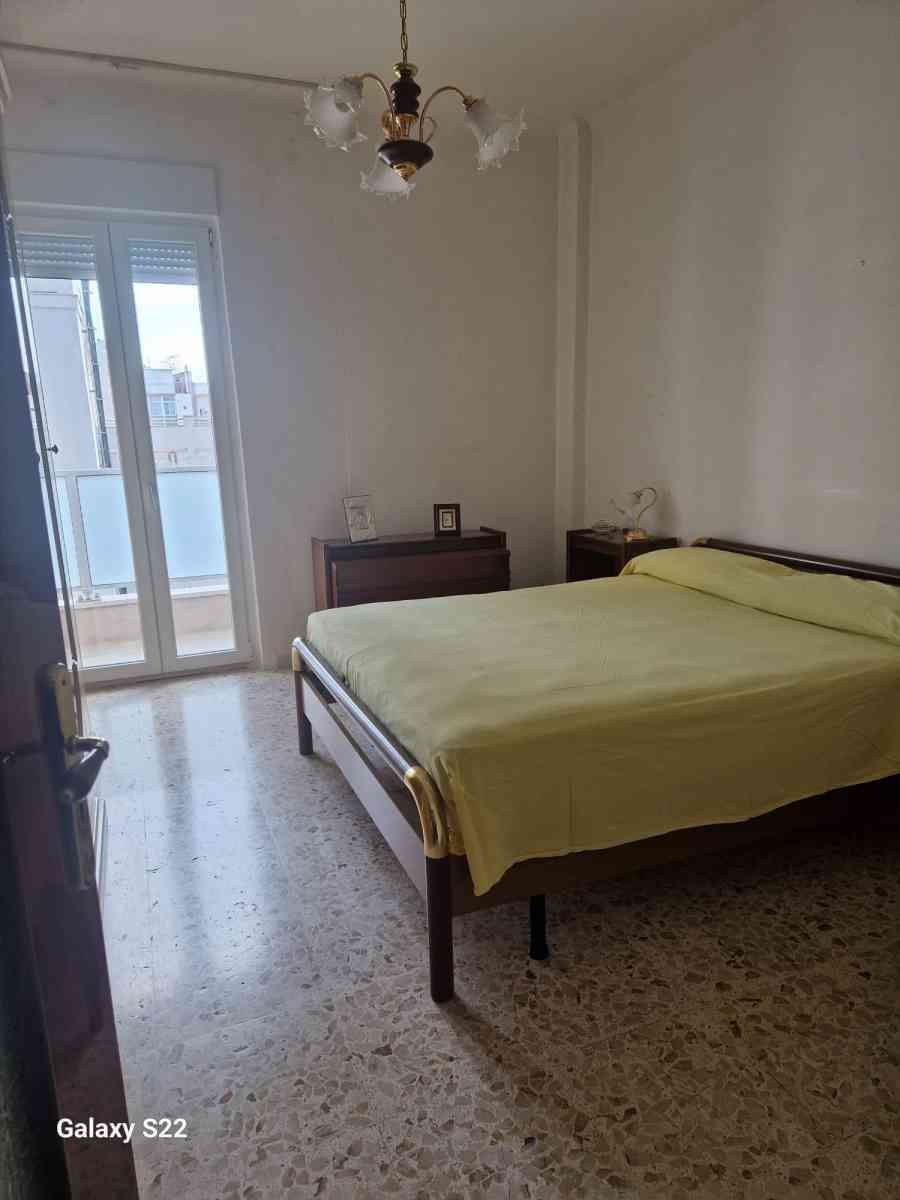 Camera degli ospiti con letto matrimoniale, comodino piccolo, pavimento piastrellato e porta finestra che lascia entrare la luce naturale.