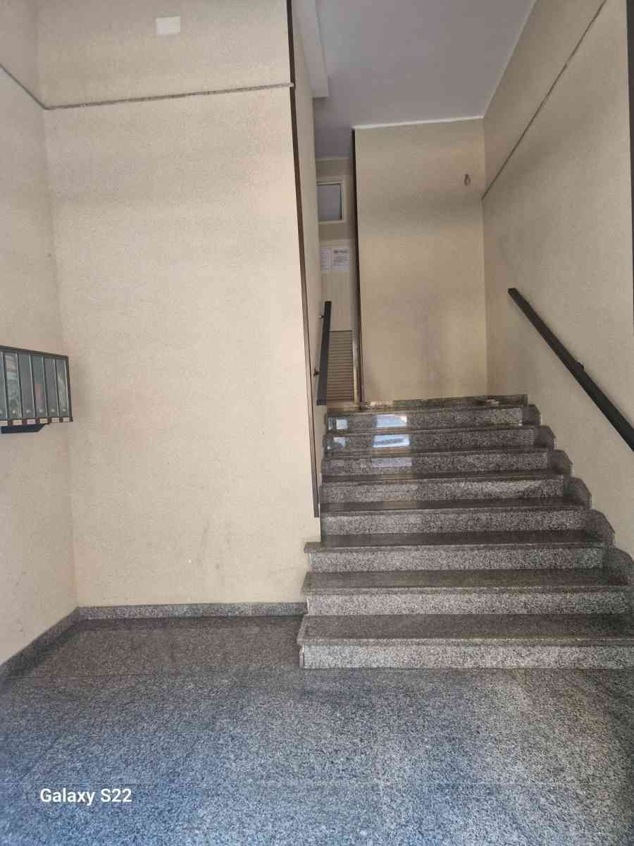 Vano scale interno con pareti beige, pavimento grigio e corrimano in metallo che conduce a un pianerottolo.