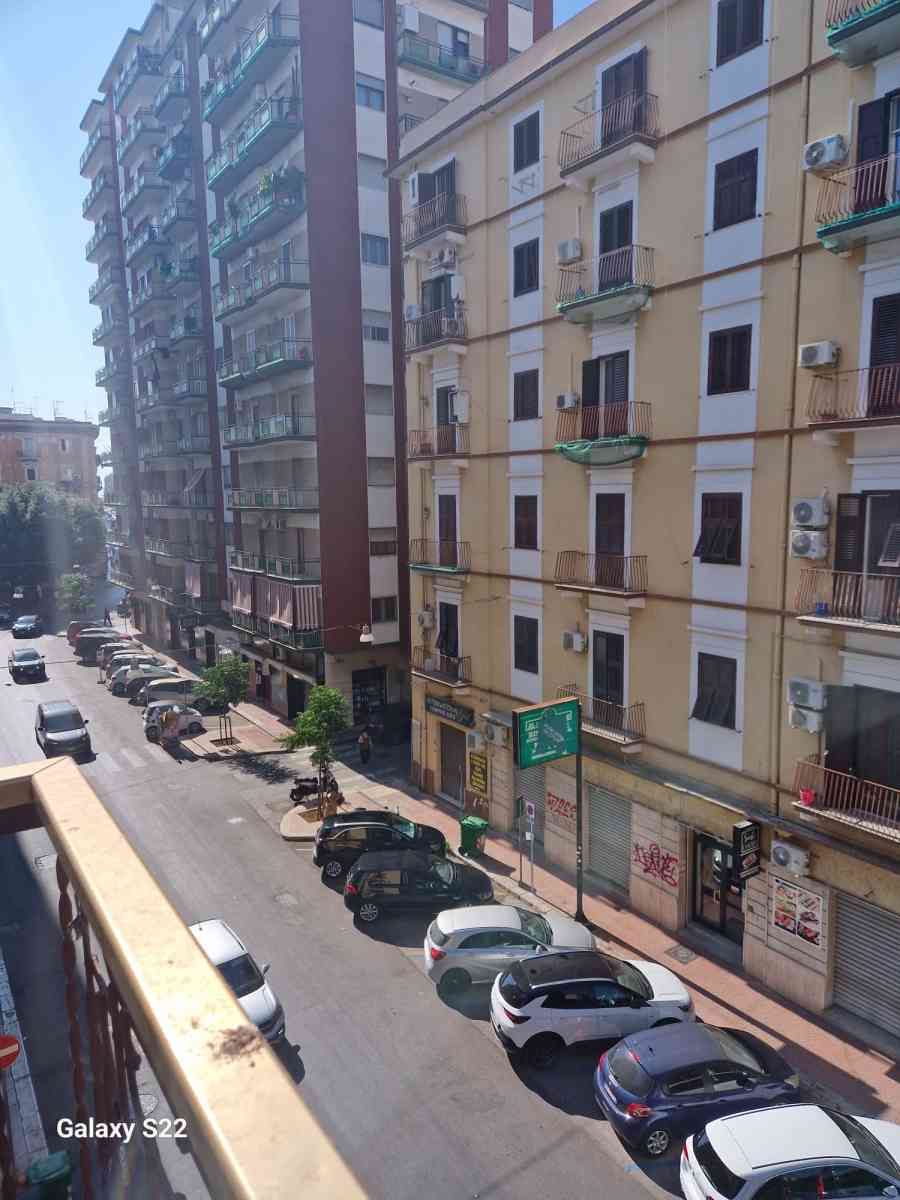 Via urbana soleggiata con auto parcheggiate, alti palazzi e balconi su un ripido pendio.