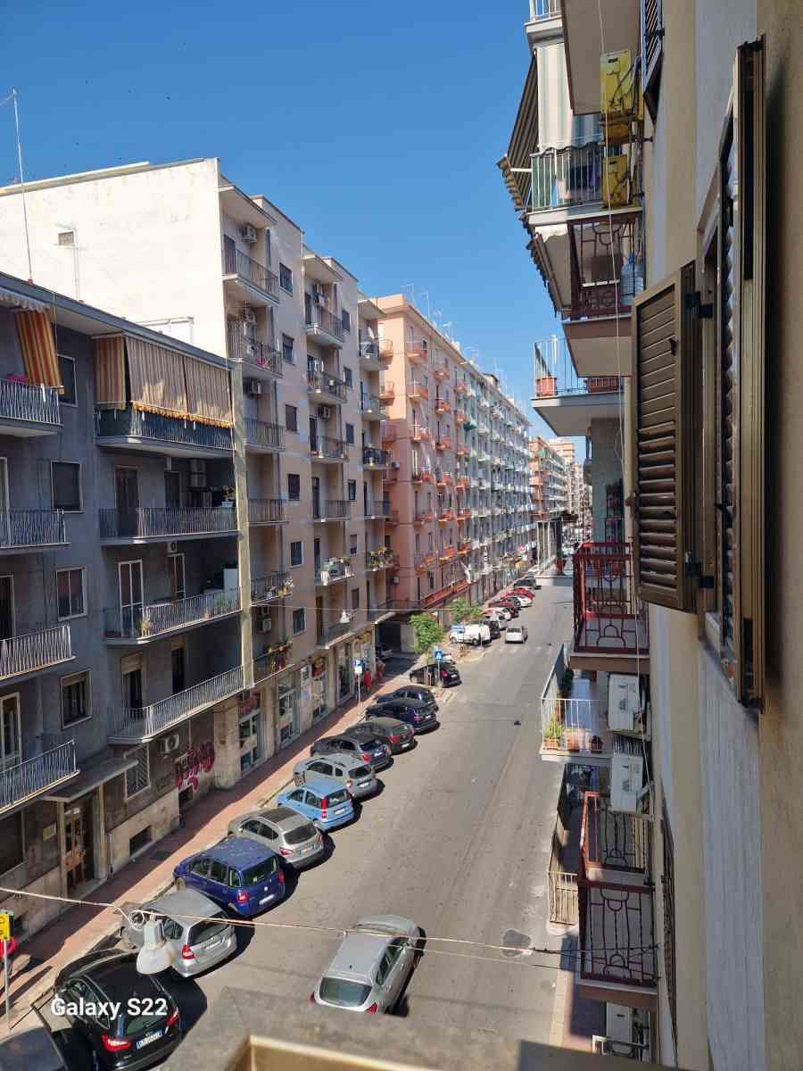 Una stretta via cittadina fiancheggiata da palazzi e auto parcheggiate sotto un cielo azzurro e limpido.