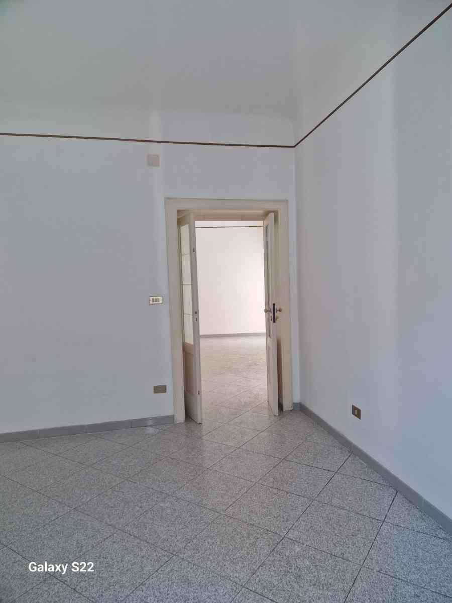 Stanza bianca vuota con pavimento piastrellato e porta aperta che conduce a un'altra stanza.