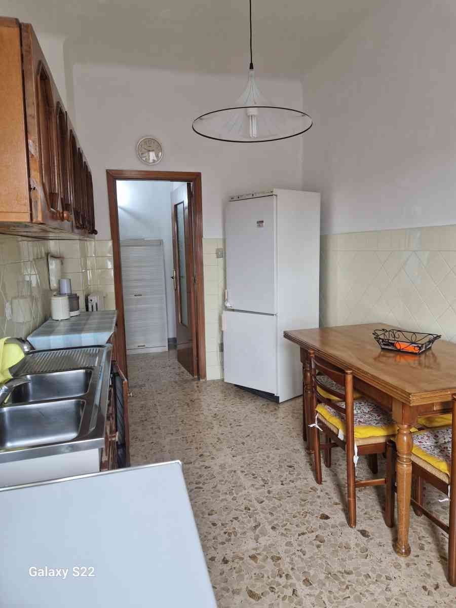 Cucina stretta con tavolo in legno, frigorifero bianco, lavello e pavimento piastrellato.