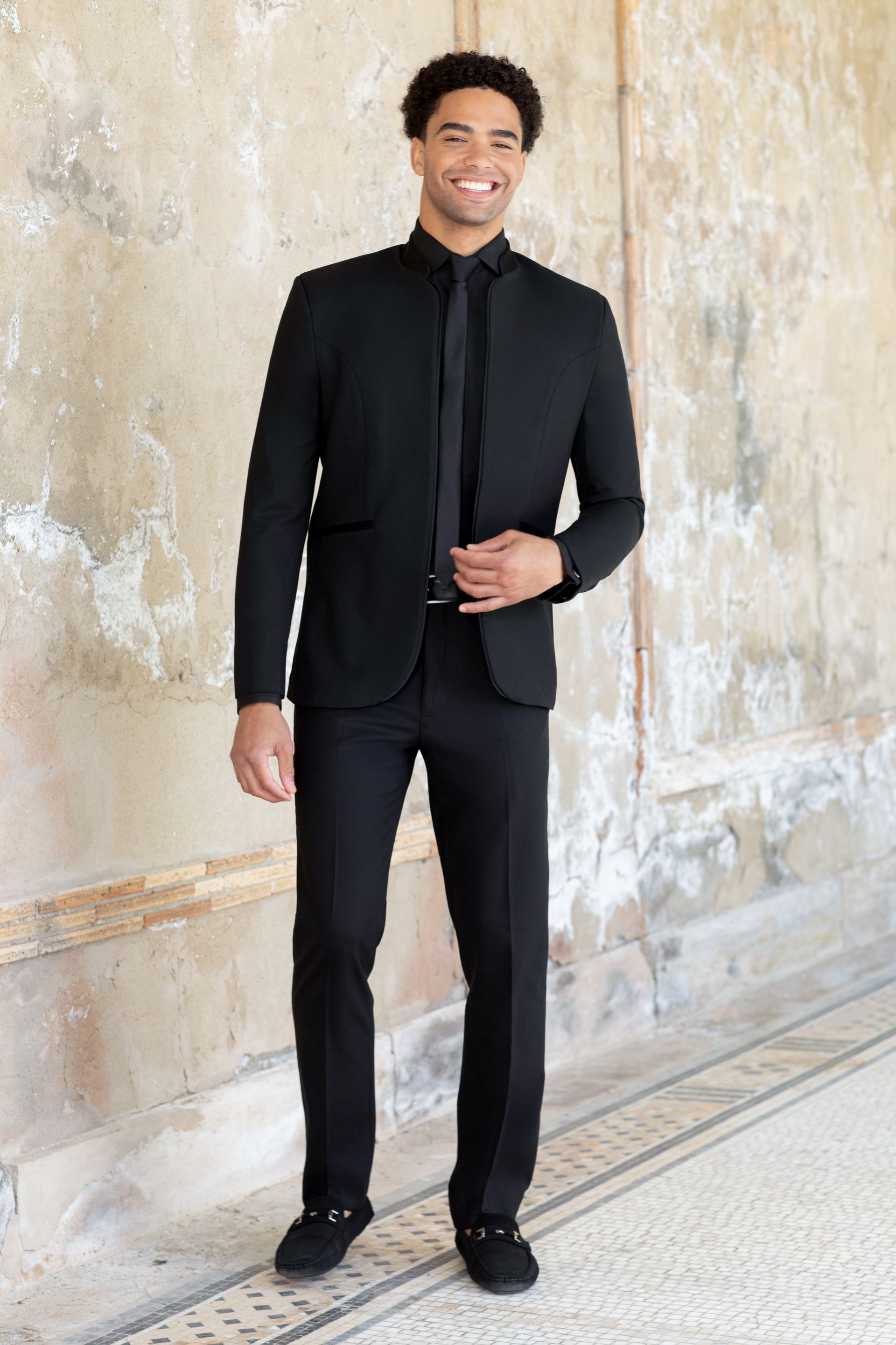 A man in a black suit and tie is standing in front of a wall.