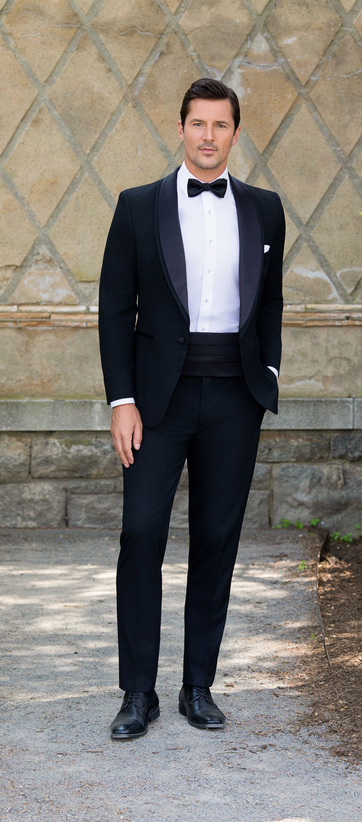 A man in a tuxedo and bow tie is standing in front of a wall.