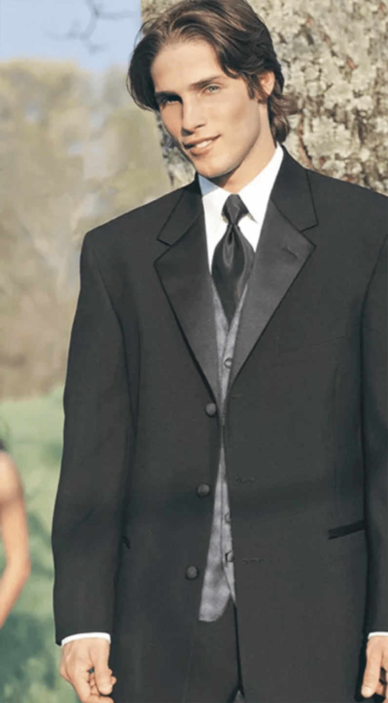 A man in a black suit and tie is leaning against a tree