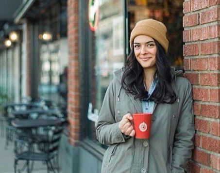 A woman is leaning against a brick wall holding a cup of coffee.