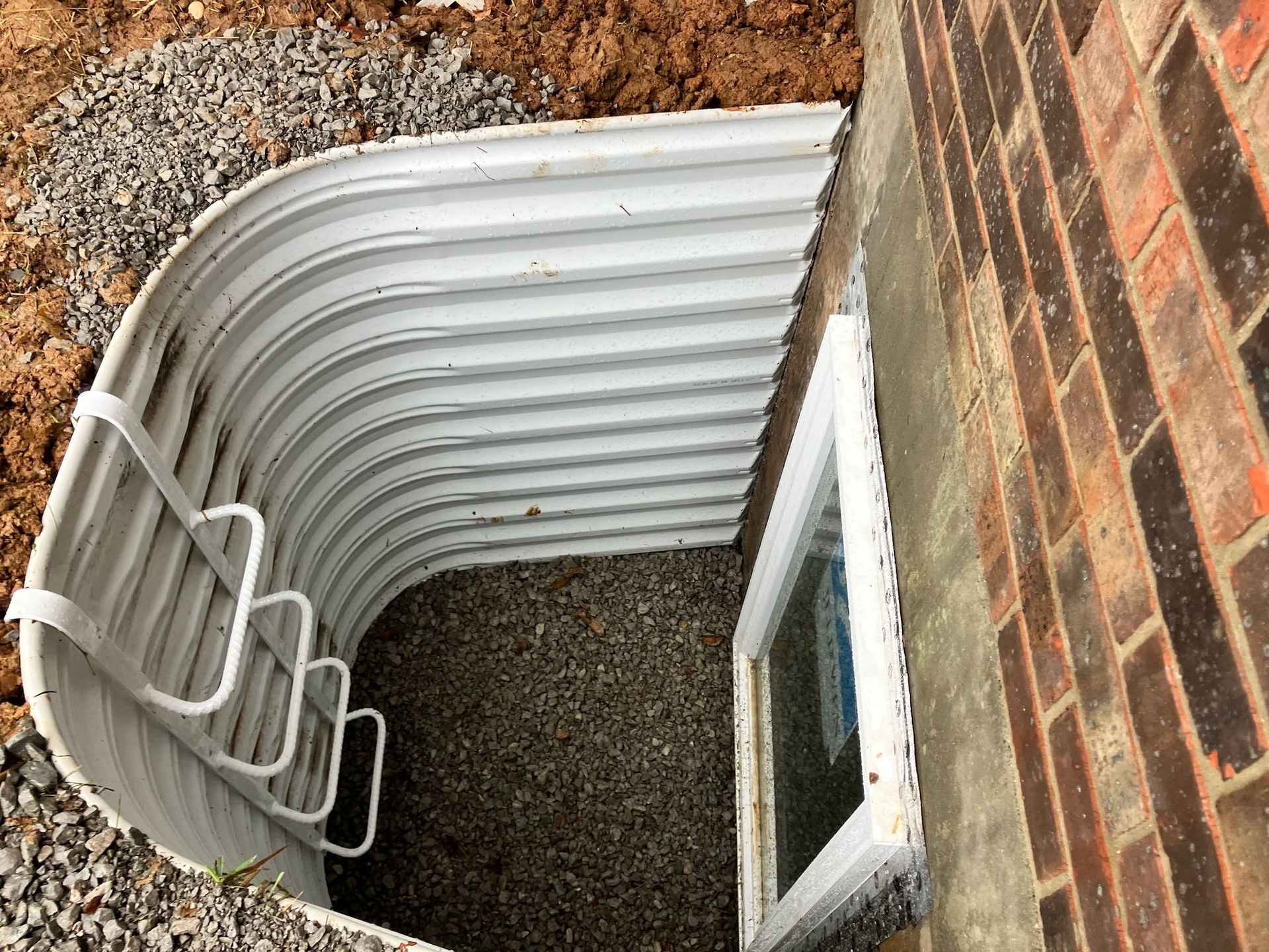 Curved, corrugated metal window well with gravel bottom next to a brick building. Includes a white egress ladder and a basement window.