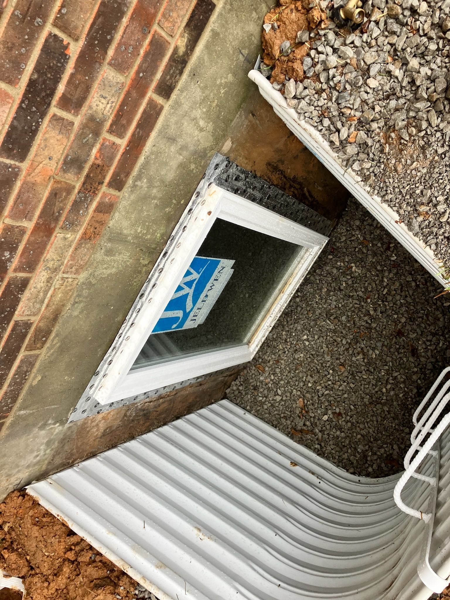 Window well with a white frame and corrugated metal sides; set in gravel next to a brick wall.