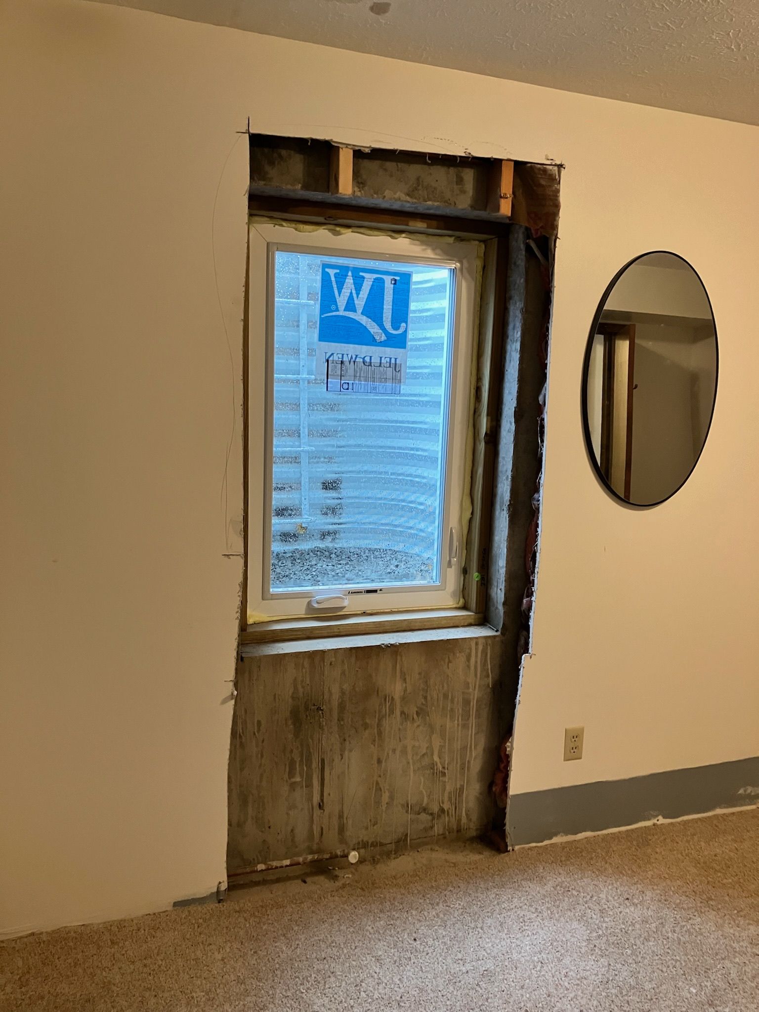 Wall with an exposed window frame; unfinished drywall; oval mirror on right.