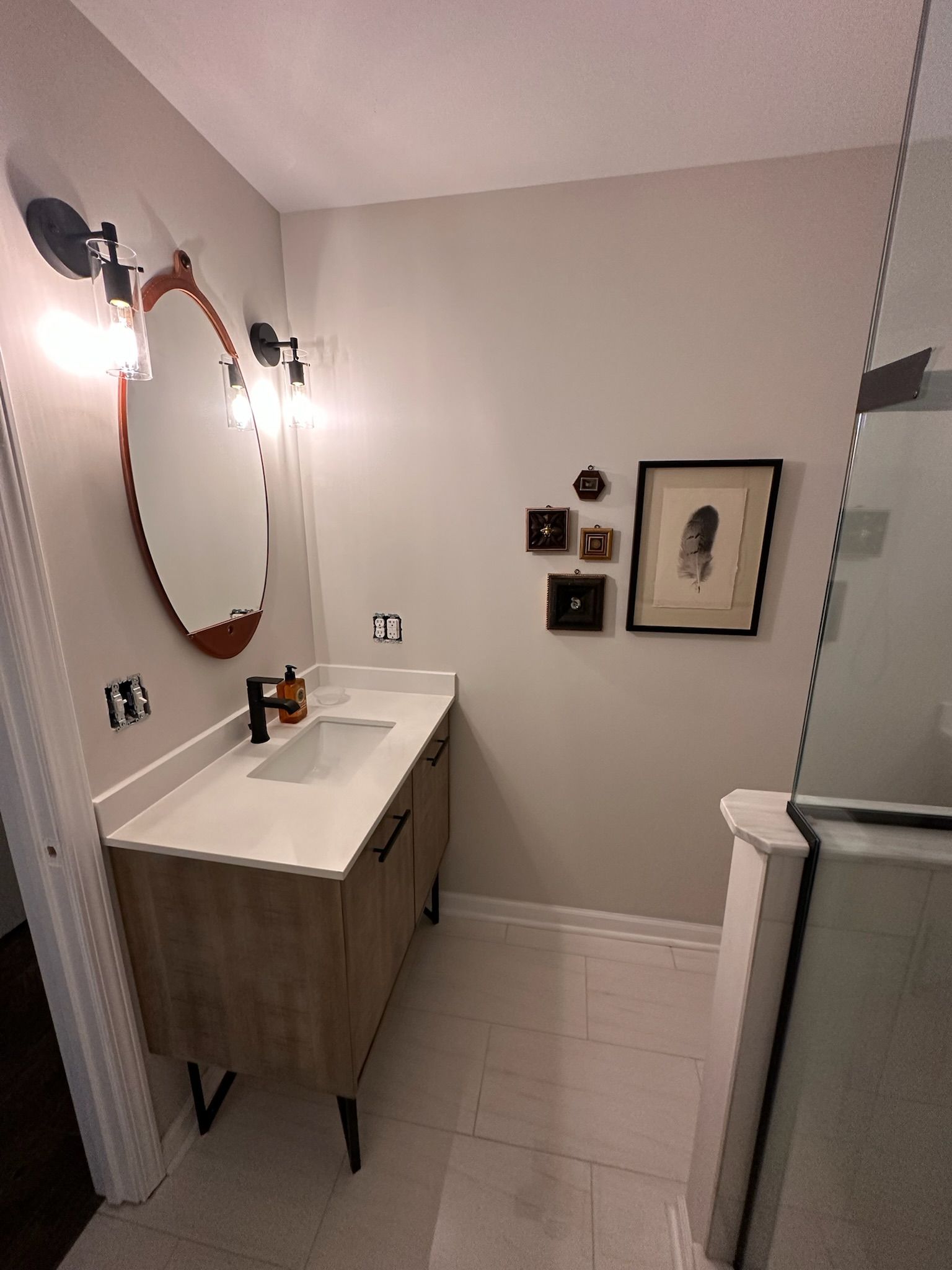 Bathroom with wooden vanity, oval mirror, sconces, and artwork on a light gray wall.