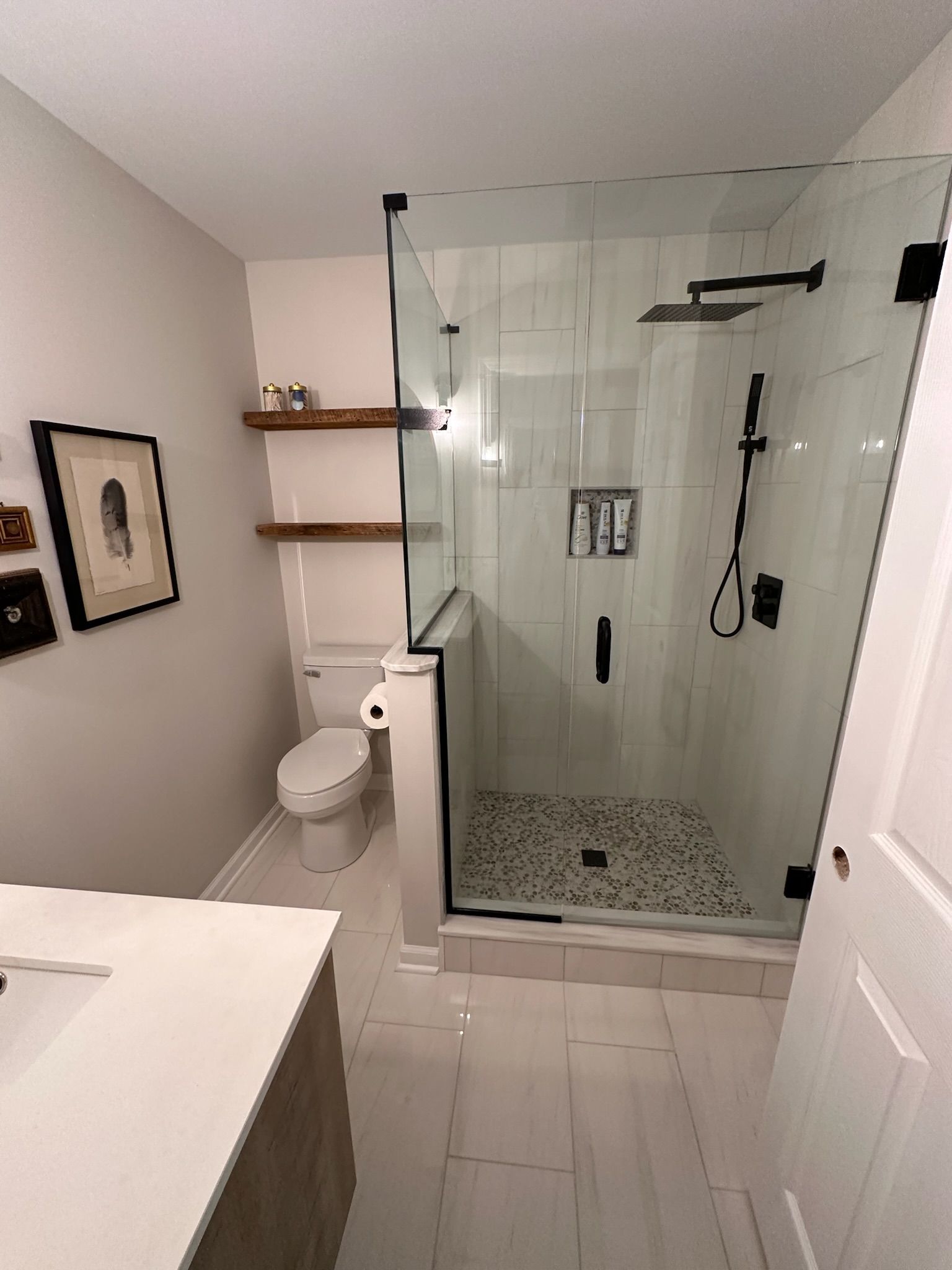 Bathroom with glass shower, white tile, black fixtures, toilet, floating shelves, and sink.