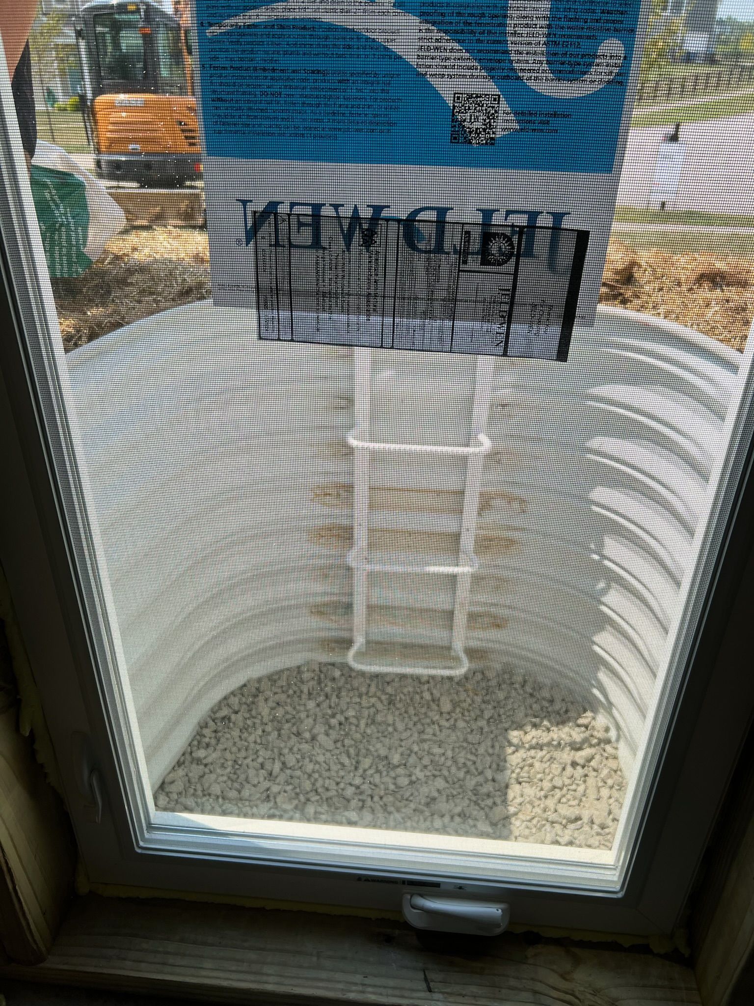 Basement window with screen, showing a light-colored window well filled with gravel. Blue sign visible through the window.