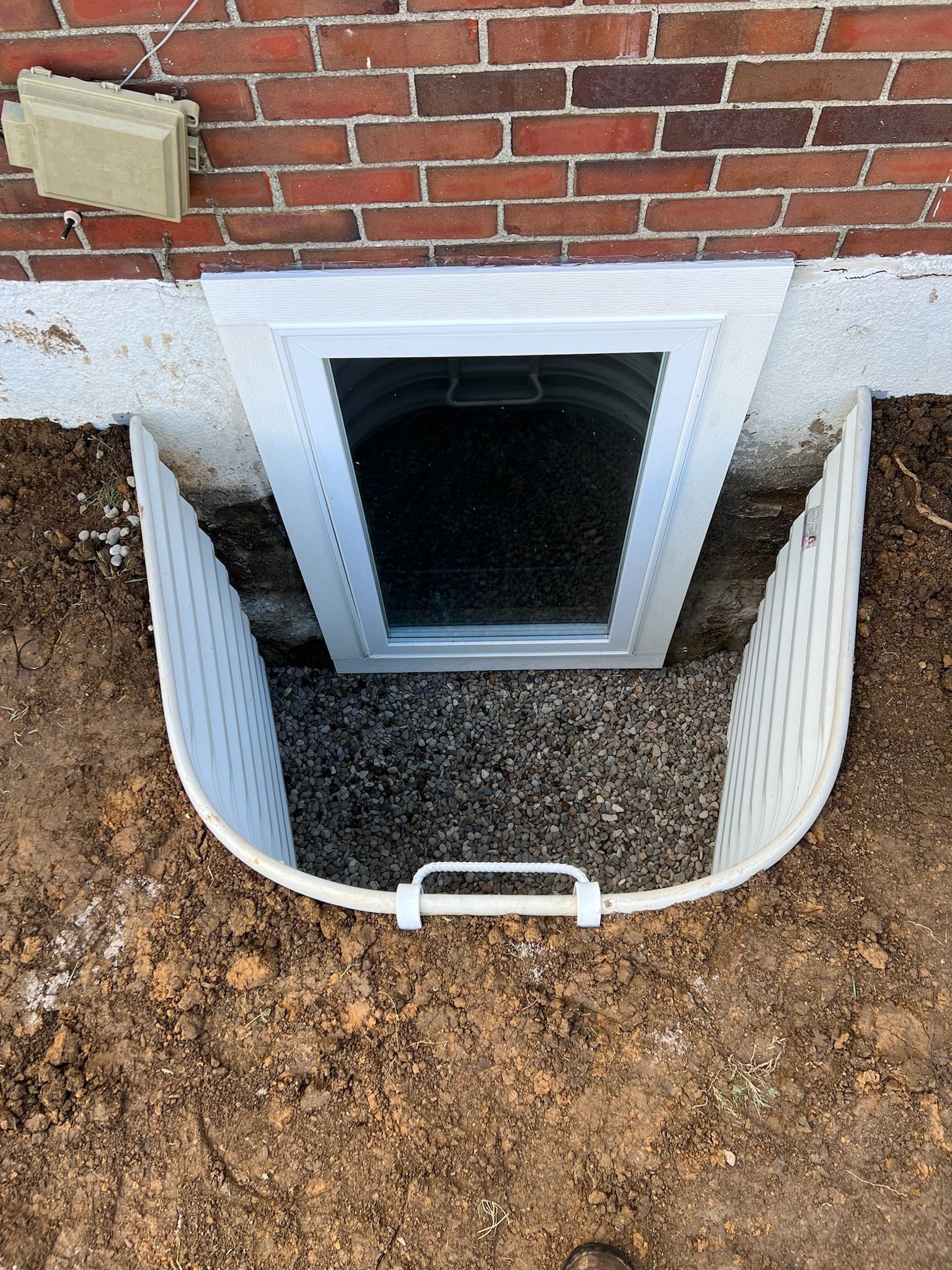 White plastic window well with a window to a basement. Gravel fills the well. Red brick wall above.