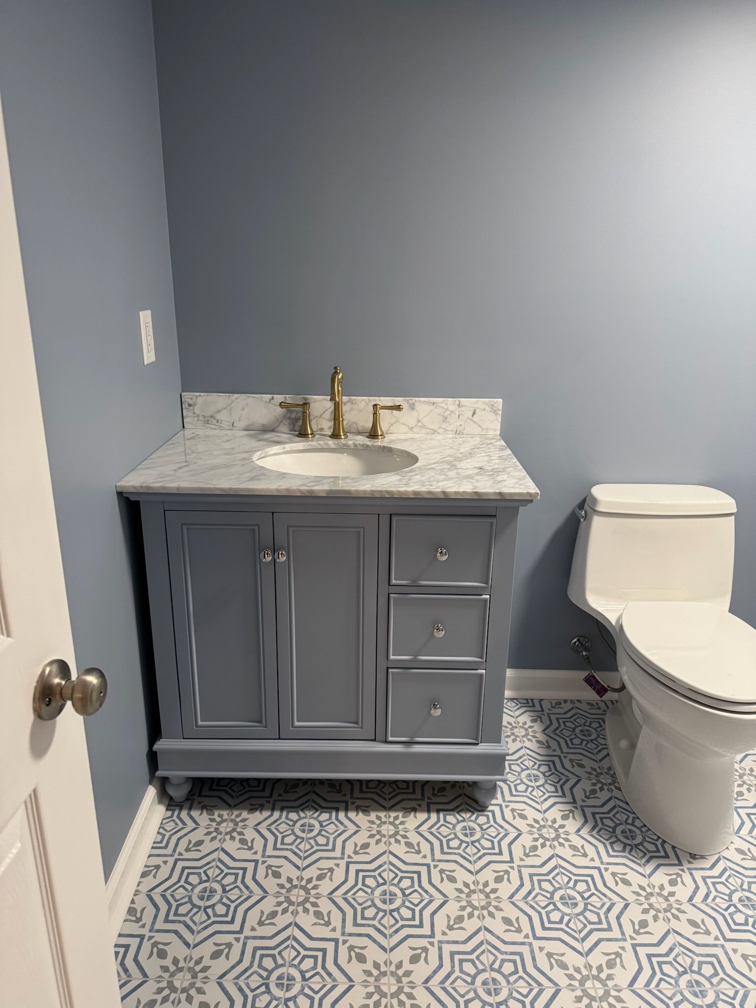 Gray vanity with marble top, gold faucet, and blue walls. Patterned floor and white toilet.
