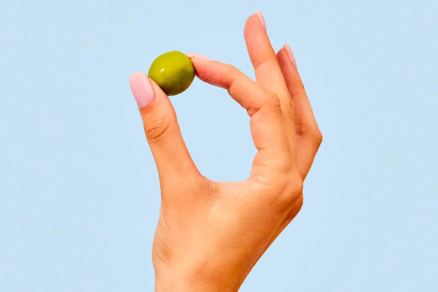 Close-up of a hand holding a fresh green olive against a light blue background, highlighting