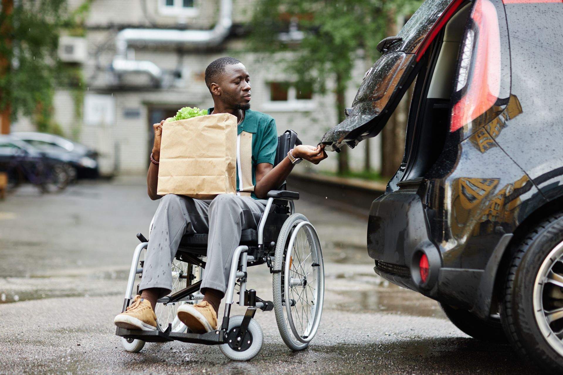 Person in a wheelchair reaches toward the open trunk of a black car on a wet street.