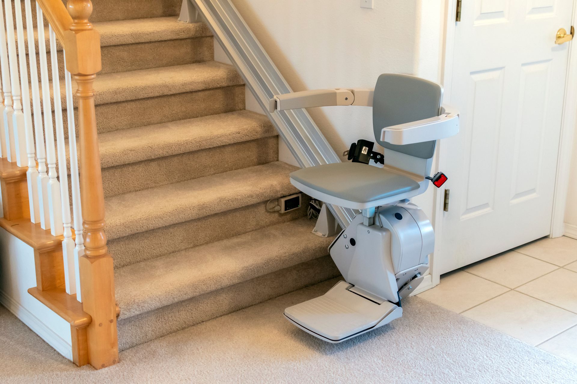 Gray stair lift beside a carpeted staircase and white door in a home hallway Gray stair lift beside a carpeted staircase and white door in a home hallway