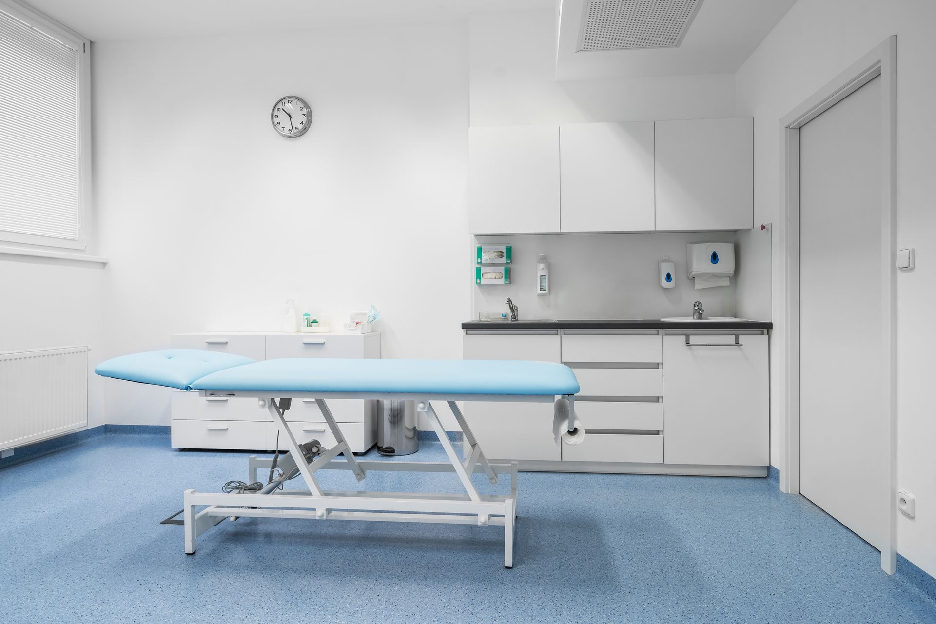 Empty medical exam room with examination table, sink, cabinets, and wall clock Empty medical exam room with examination table, sink, cabinets, and wall clock