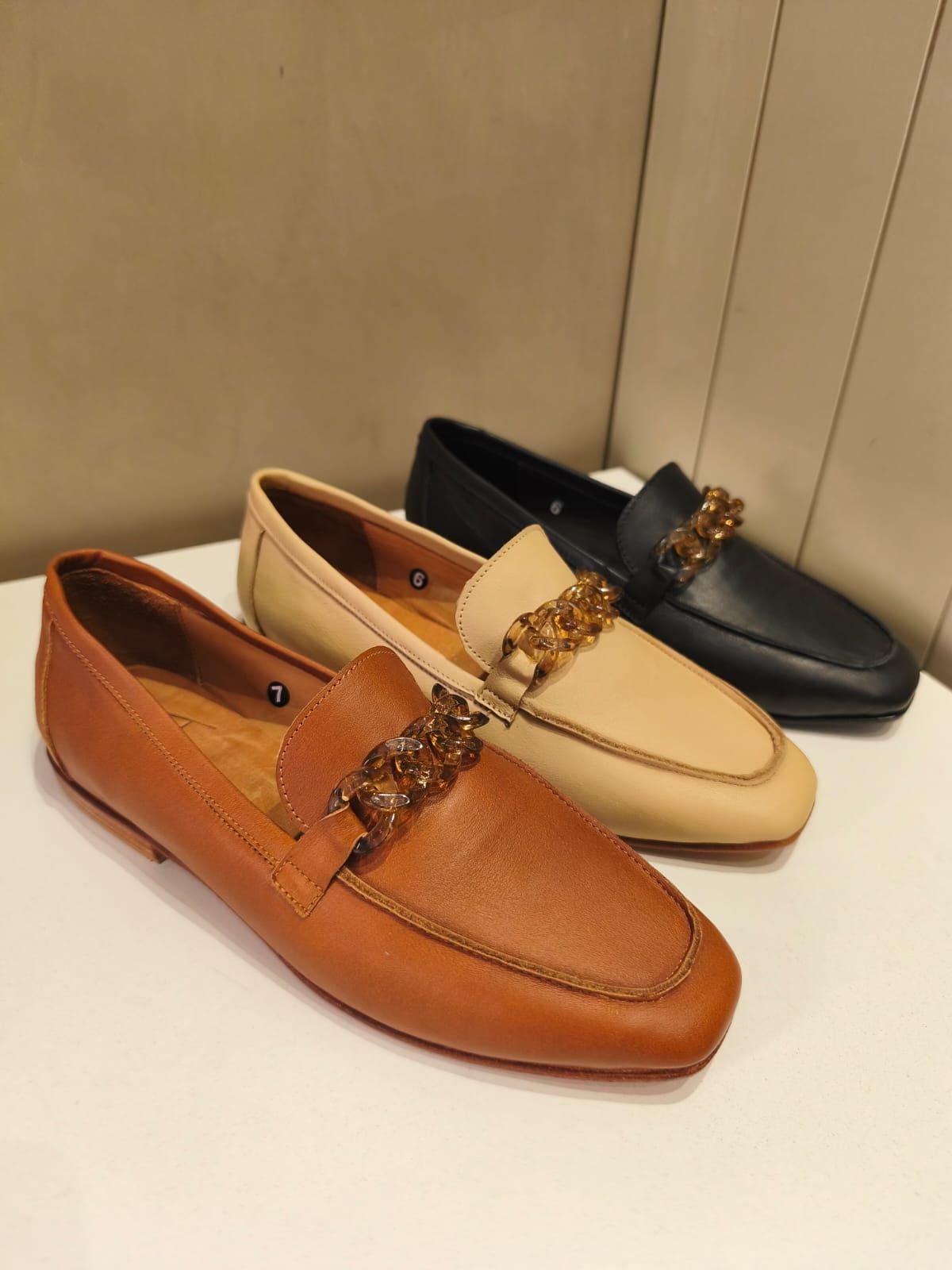 Tres mocasines de piel en color marrón, beige y negro con detalle de cadena dorada.