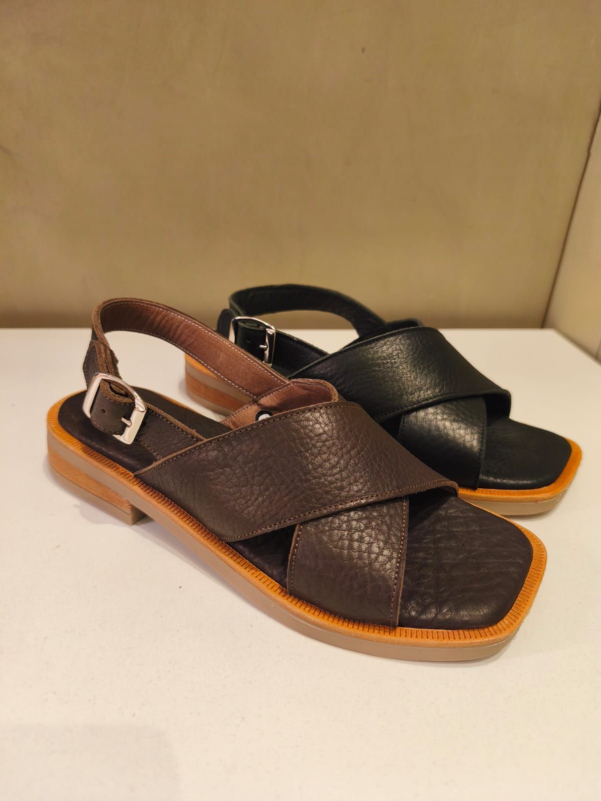 Dos sandalias de piel, una marrón y otra negra, con tiras cruzadas y hebillas en el tobillo.