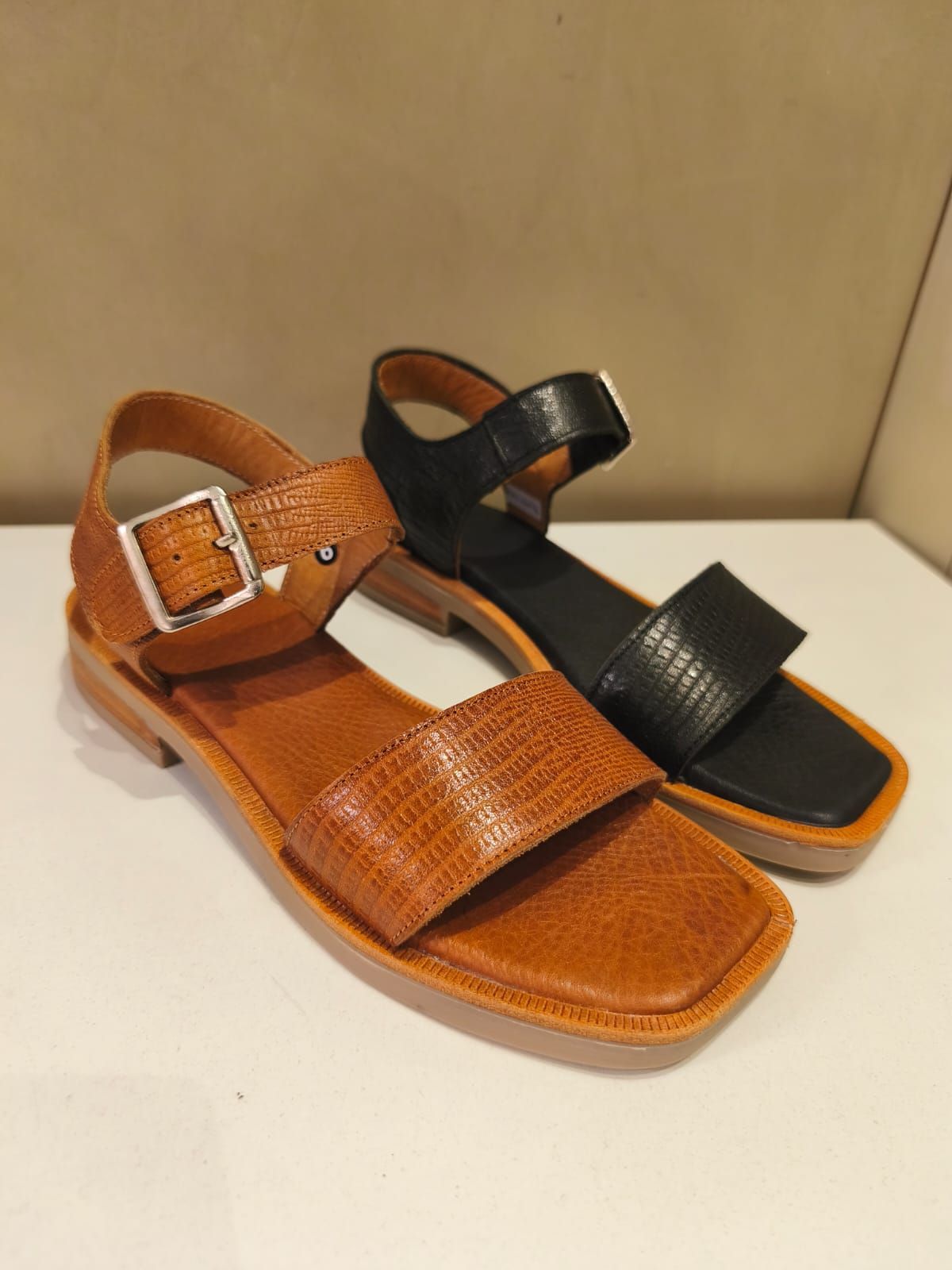 Dos sandalias, una marrón y otra negra, expuestas sobre una superficie blanca.