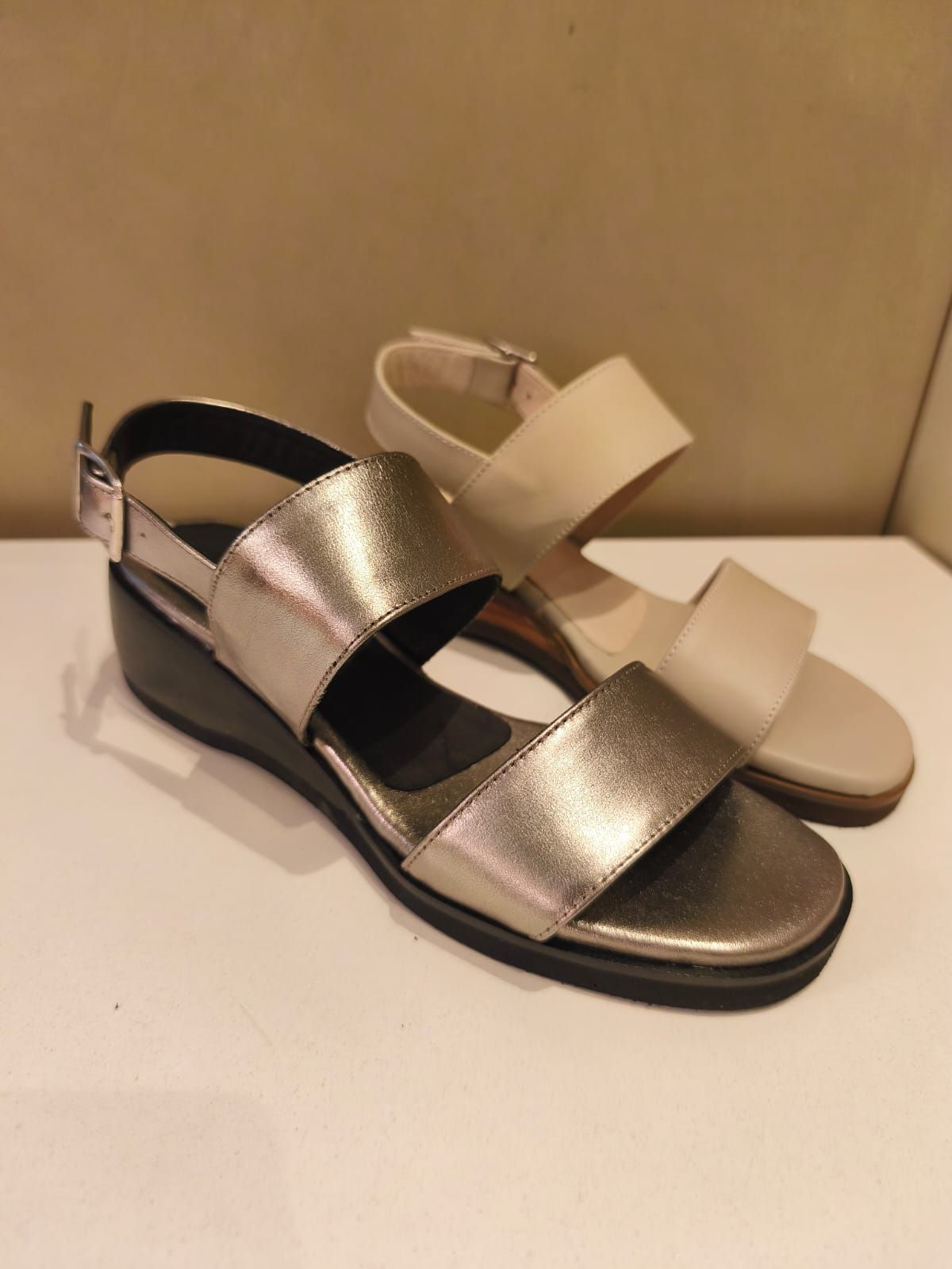 Dos pares de sandalias de mujer sobre una superficie blanca. Un par es plateado metalizado y el otro beige.