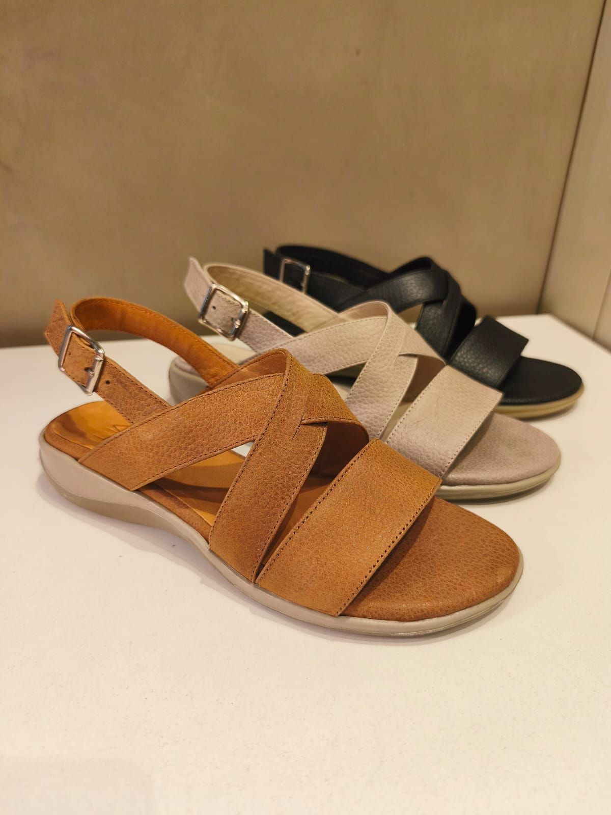 Tres pares de sandalias de mujer en marrón, beige y negro con tiras entrecruzadas, sobre una superficie blanca.