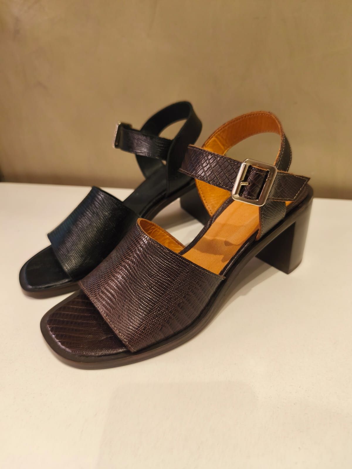 Dos sandalias de tacón bloque, una negra y otra marrón, con tiras en el tobillo, sobre una superficie blanca.
