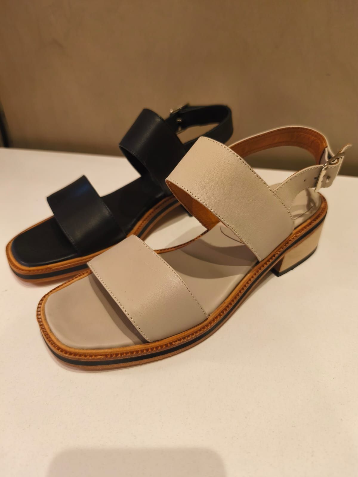 Dos sandalias de piel, una negra y otra beige, expuestas sobre una superficie blanca.