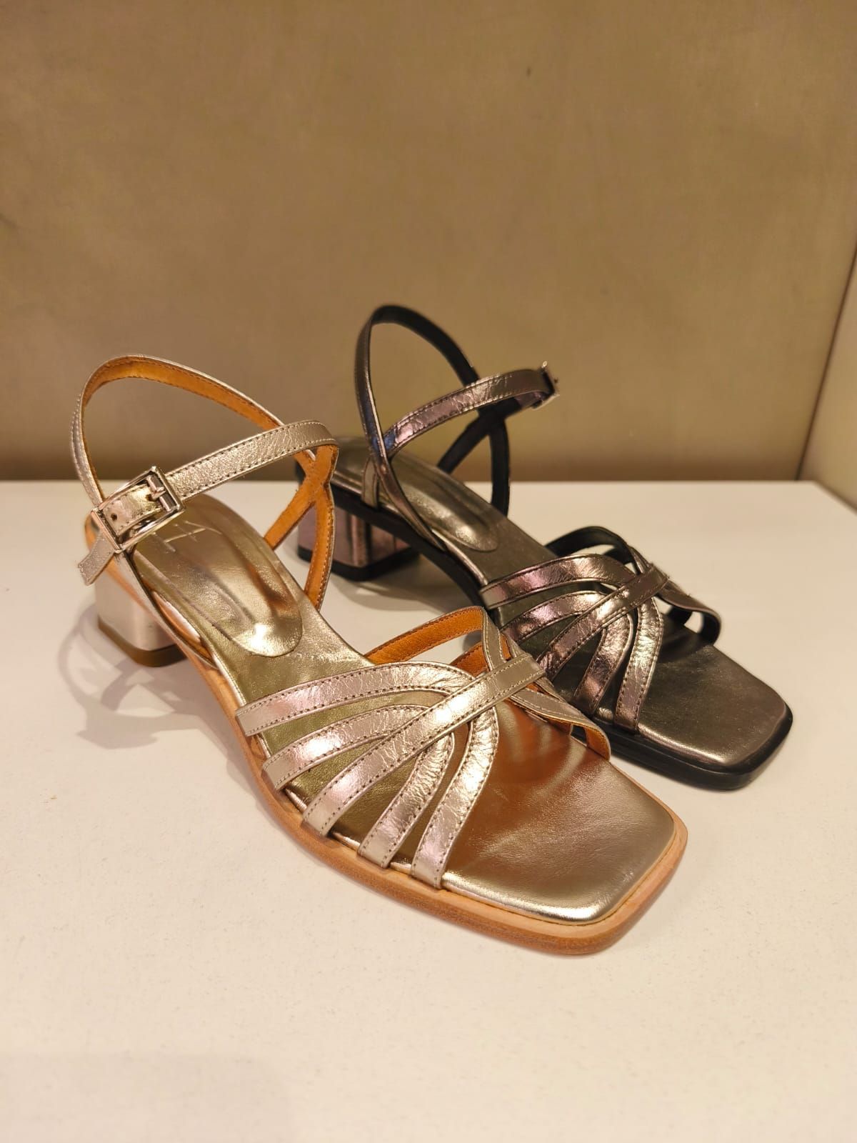 Dos sandalias metálicas de tiras: doradas y negras. Punta cuadrada y tacón bajo. Sobre una superficie blanca.