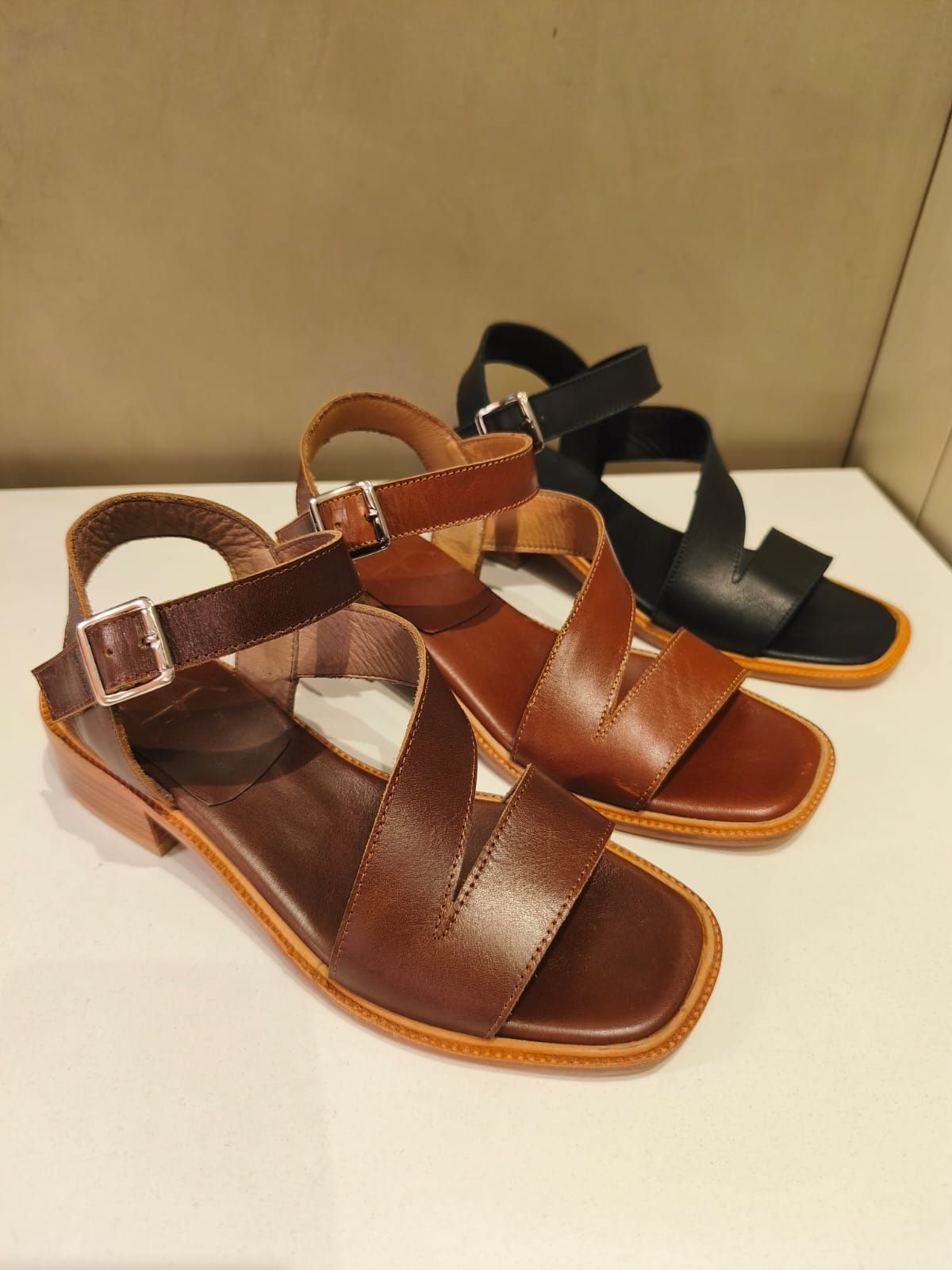 Tres sandalias de cuero en color marrón, tostado y negro sobre una superficie blanca.