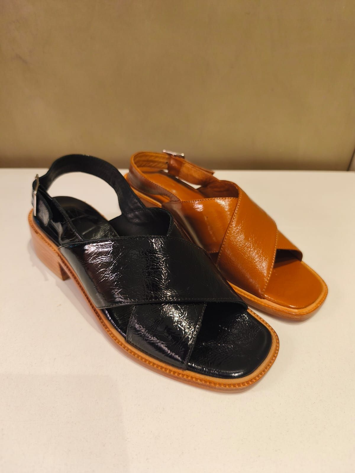 Dos sandalias de mujer, una negra y otra marrón, se encuentran una al lado de la otra sobre una superficie blanca.