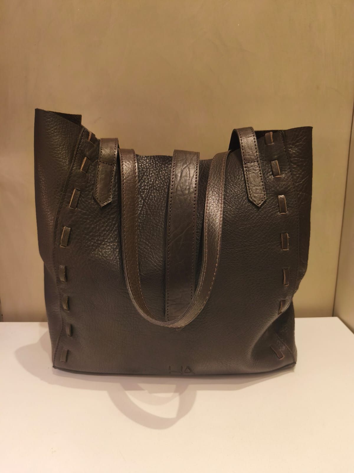 Bolso de mano de piel de color marrón oscuro con asas, sobre una superficie blanca sobre un fondo tostado.