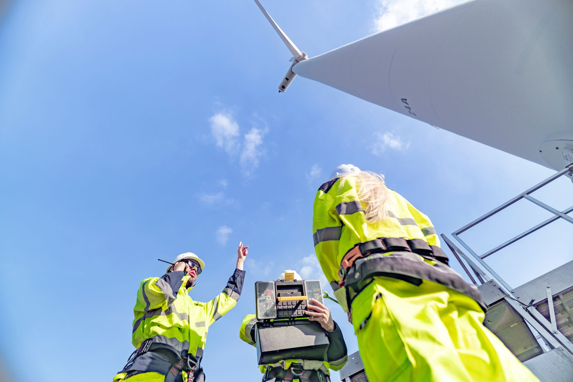 Workers in yellow safety vests inspect a wind turbine blade, under a blue sky. One worker points upward. Workers in yellow safety vests inspect a wind turbine blade, under a blue sky. One worker points upward.