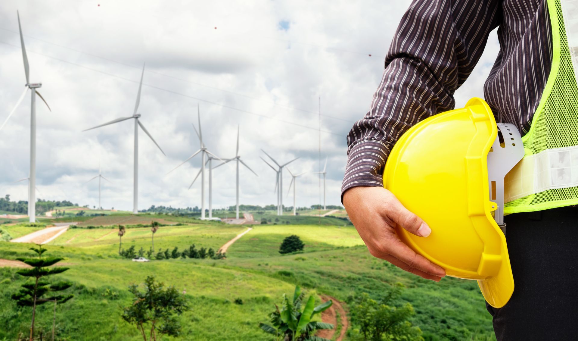 Engineer holding a yellow hard hat, standing in front of a wind farm on a green, grassy hill. Engineer holding a yellow hard hat, standing in front of a wind farm on a green, grassy hill.